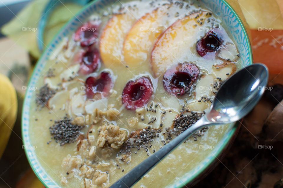 Beautiful fresh fruit smoothie bowl ready to eat with spoon on rim of bowl garnished with black cherry, sliced peach, oatmeal, coconut, chia seed and sliced almonds part of a series 