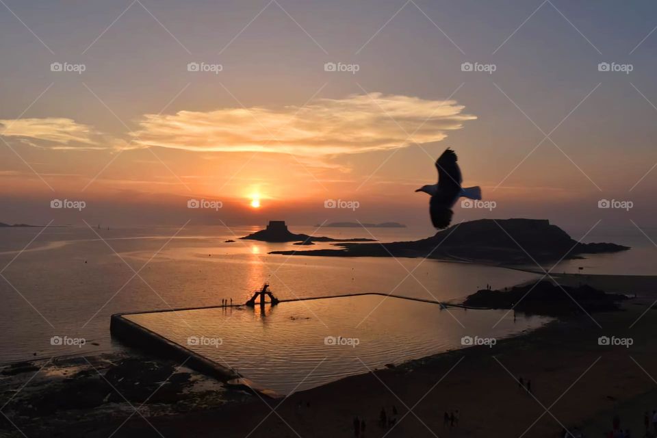 View of the Bon Secours beach in Saint Malo showing its diving metallic platform and a bird flying by at sunset under a grey and orange sky
