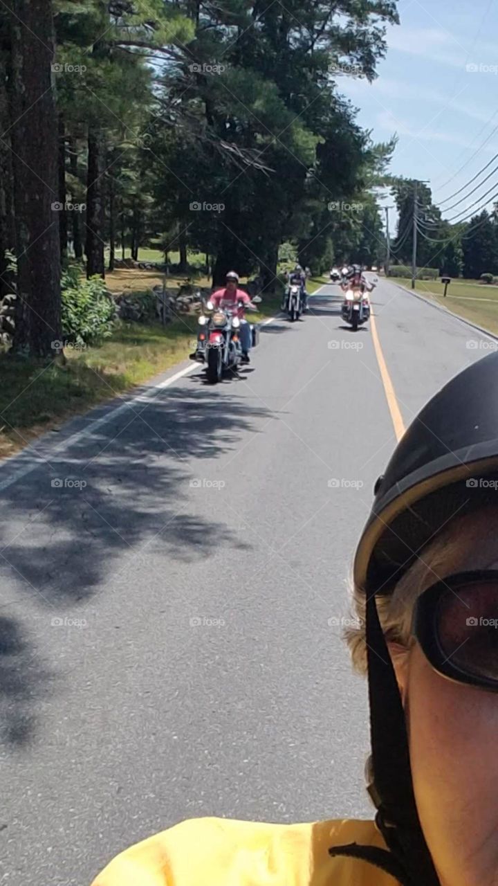 Road in rural America looking over shoulder from motorcycle back seat. Several riders on road behind passenger also on motorcycle. Sunny day on back road.