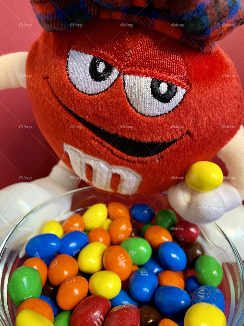 A red m&m plush toy holding a yellow m&m. There is a glass bowl with m&m 