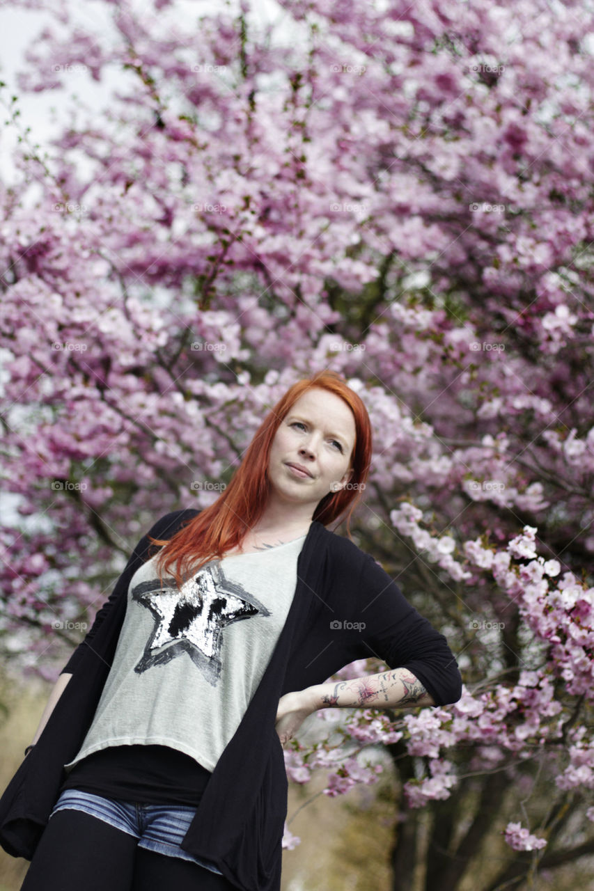A tree of cherry blossom and in front stay a woman with red hair 
