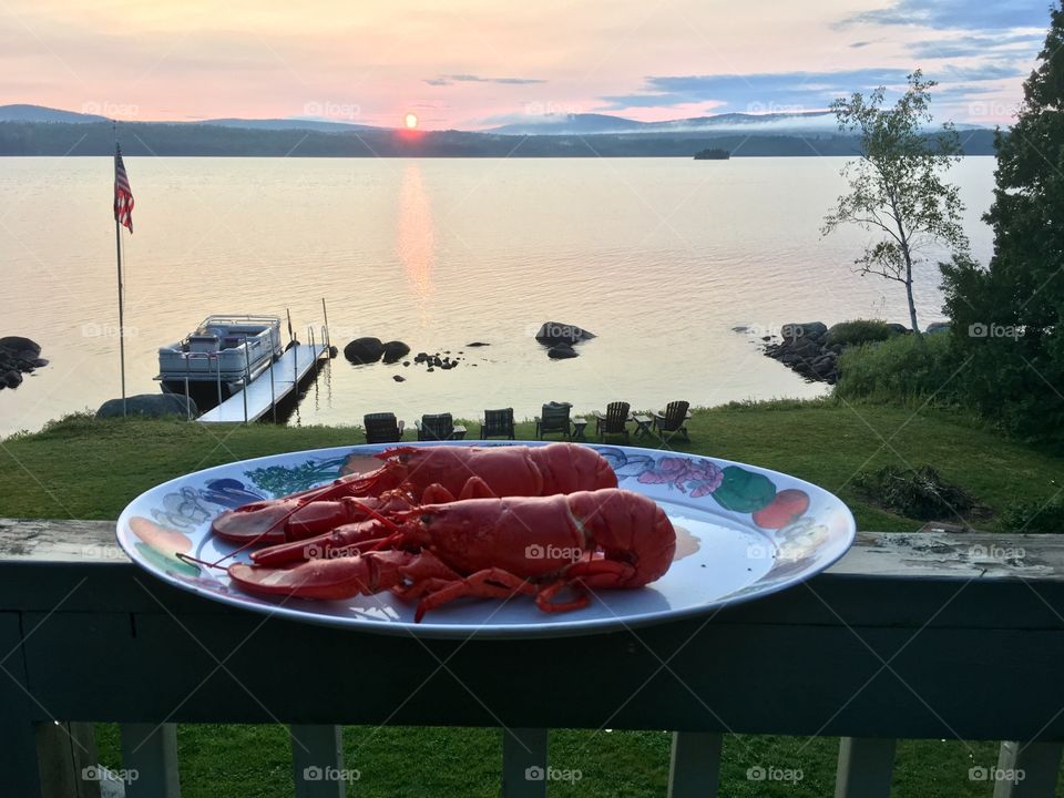 Two cooked lobsters on plate foreground with lake and boat in background at dusk