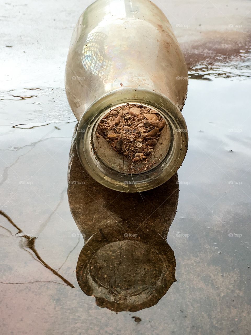 Antique milk bottle on its side in the rain and rainwater filled with sand and seashells as found and reflection in the rainwater