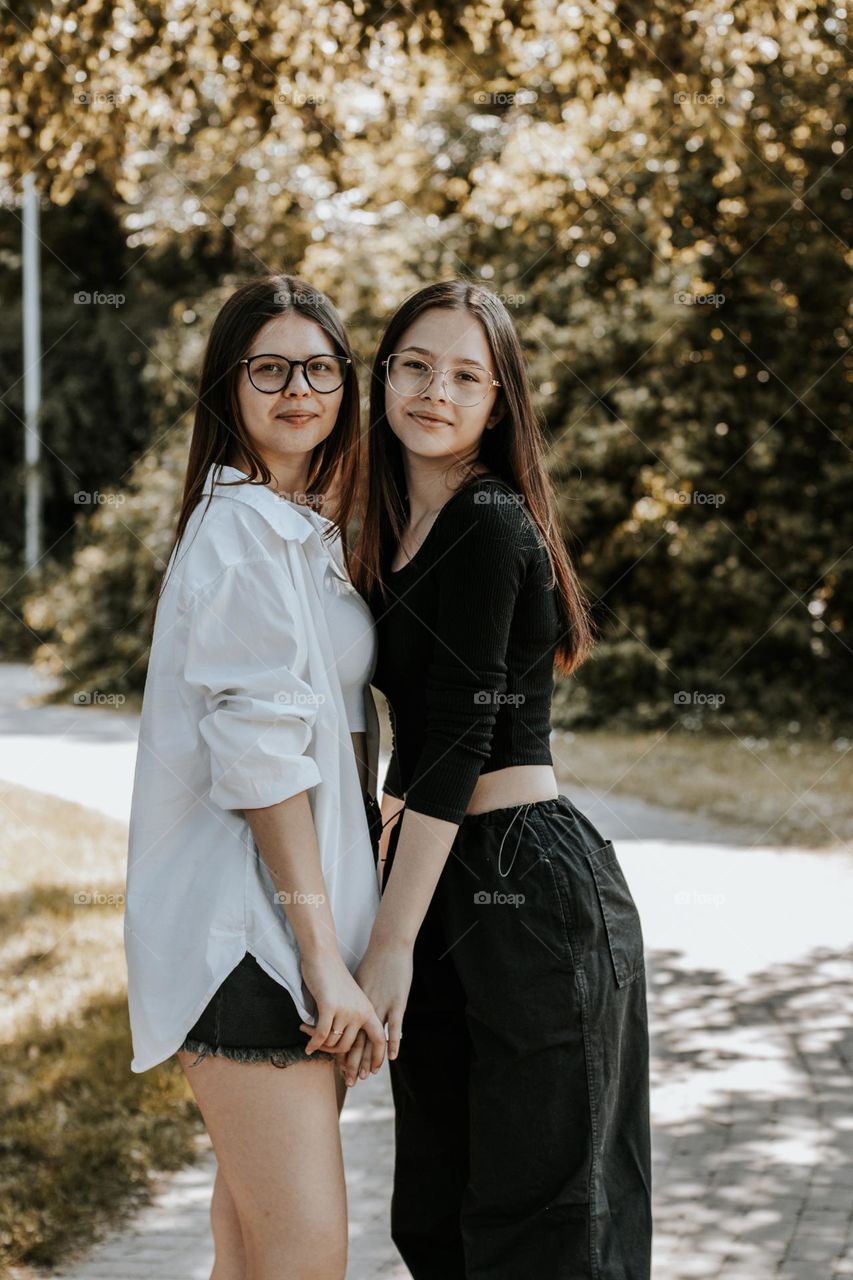 Portrait of two beautiful young Caucasian brunette girls with loose hair standing in an embrace and holding each other's hands in a public park on a summer day, close-up side view.