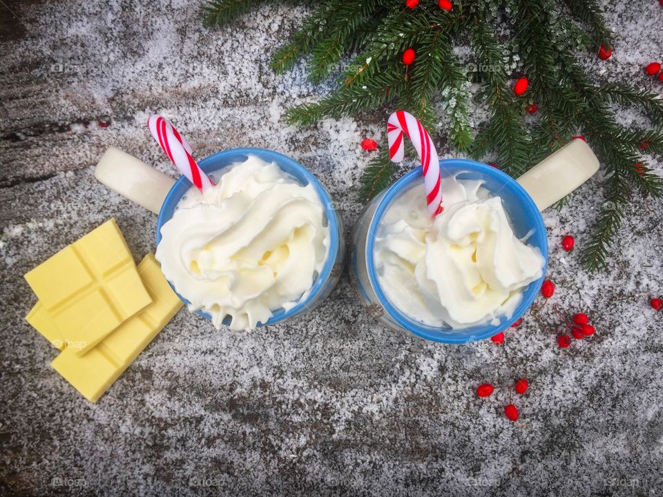 Two mugs of white chocolate with whipped cream and candy canes on a snowy table with pieces of white chocolate and evergreen branches beside