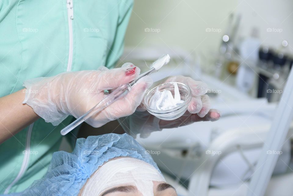 Doctor cosmetologist makes a procedure with a cream mask to a woman in a beauty salon. She wears gloves and a mask.