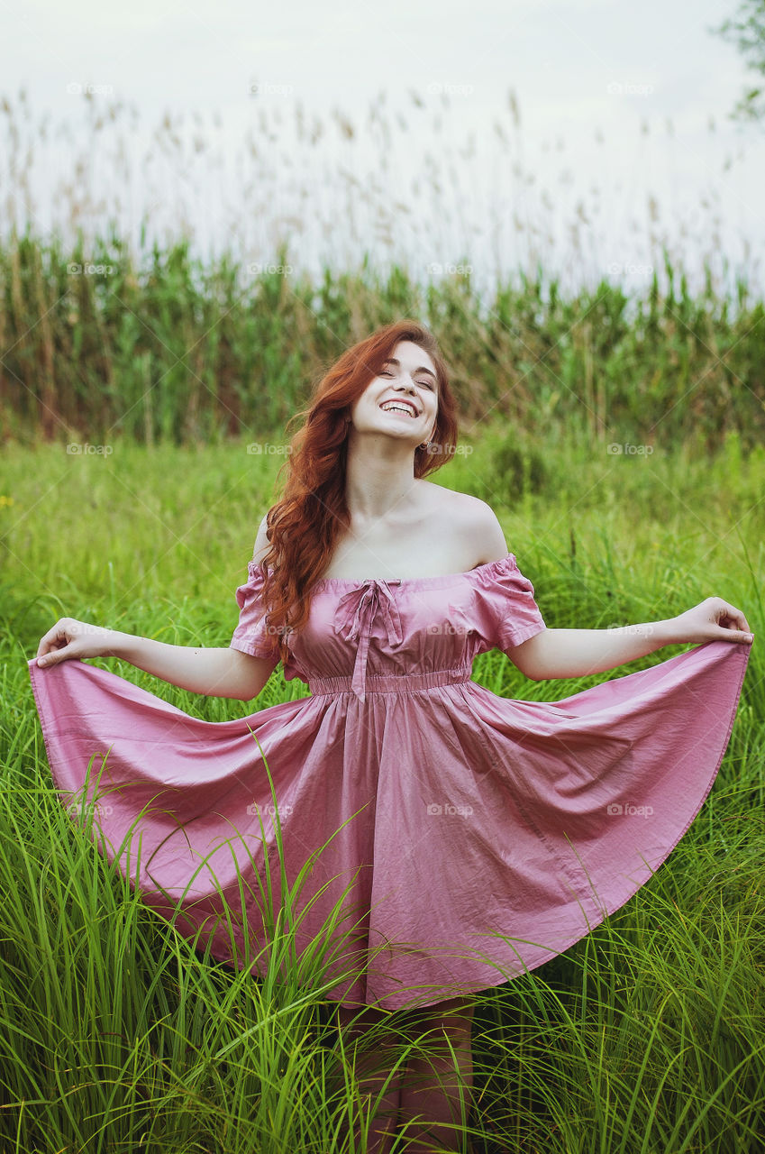 Beautiful cute redhear curly woman in pink dress dancing and smiling in the green field at summer in sunny day.