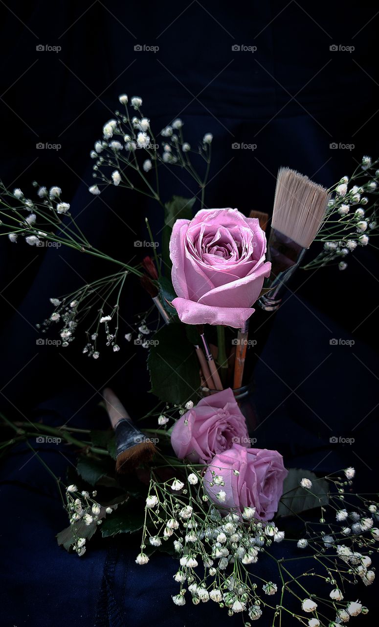A purple rose stands with brushes for paints and white small flowers. Two purple roses lie next to a paint brush and with white small flowers. Background dark blue