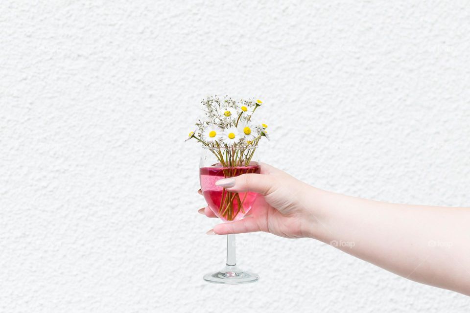 Hand holding a glass filled with red beverage and a daisy flower bouquet with white background 