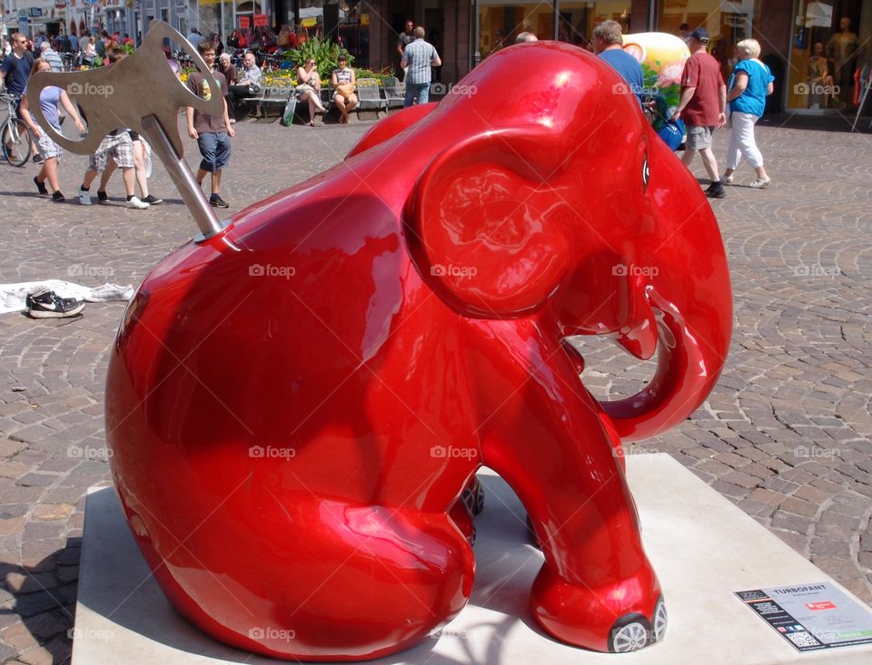 A large elephant statue with bright cherry red color and a wind up design on it show the talent of an artist in a public square in Northwestern Germany on a sunny summer day. 