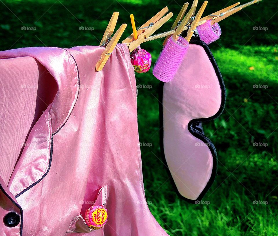 Barbie style. On a rope, with the help of wooden clothespins hang: pink pajamas, pink eye mask, pink curlers, "Chupa Chyps". Green grass in the background