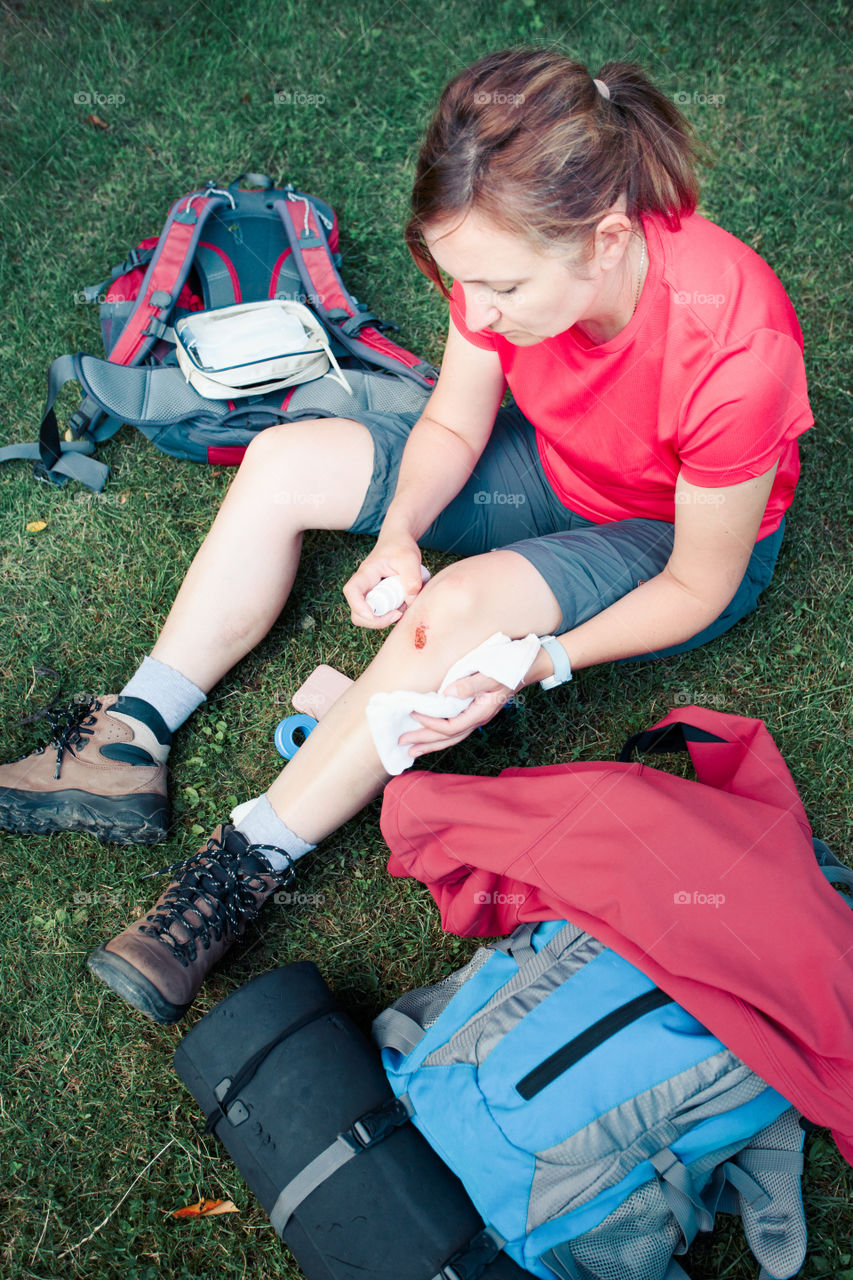 Woman injured in the accident on mountain hike. Dressing the wound on her knee with medicine in spray and gauze sitting on grass. Equipped with backpack wearing sportswear