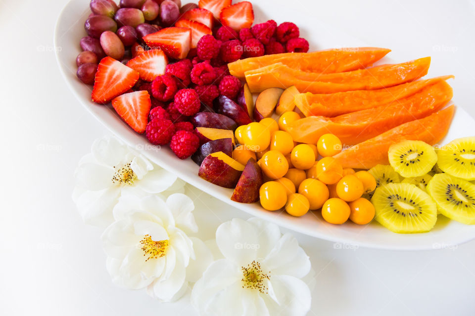 Fresh summer fruit platter with orange
, red, purple and yellow fruits with white flowers on white background. Close up fruits, different variety. Healthy fruit platter for spring and summer days.