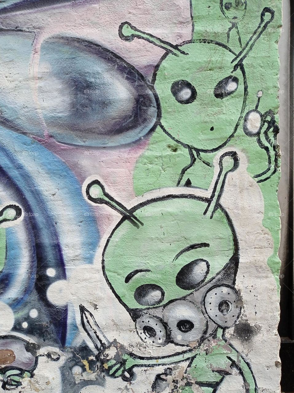 What are ETs doing? Amusing graffiti on a wall shows two green men, one looking one way with a lazer gun, one with a knife looking the other way. Friends or foes? "Defendents of the world come forth and unite."