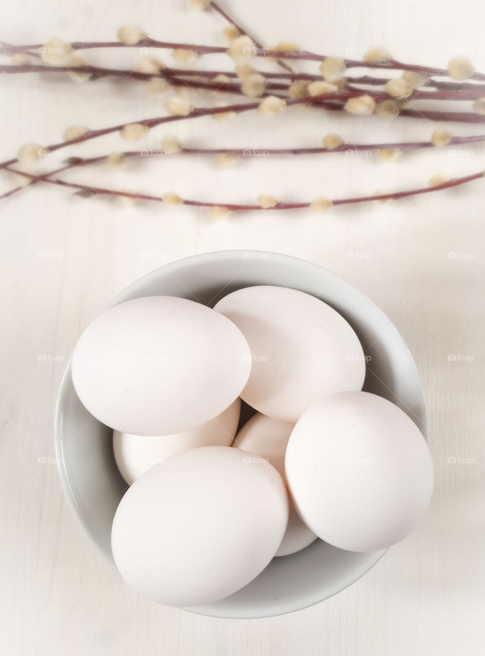 Still life with white whole raw eggs in a bowl on a white wooden table top with twigs.  Preparing for the Easter holidays.  Food photo, vertical orientation