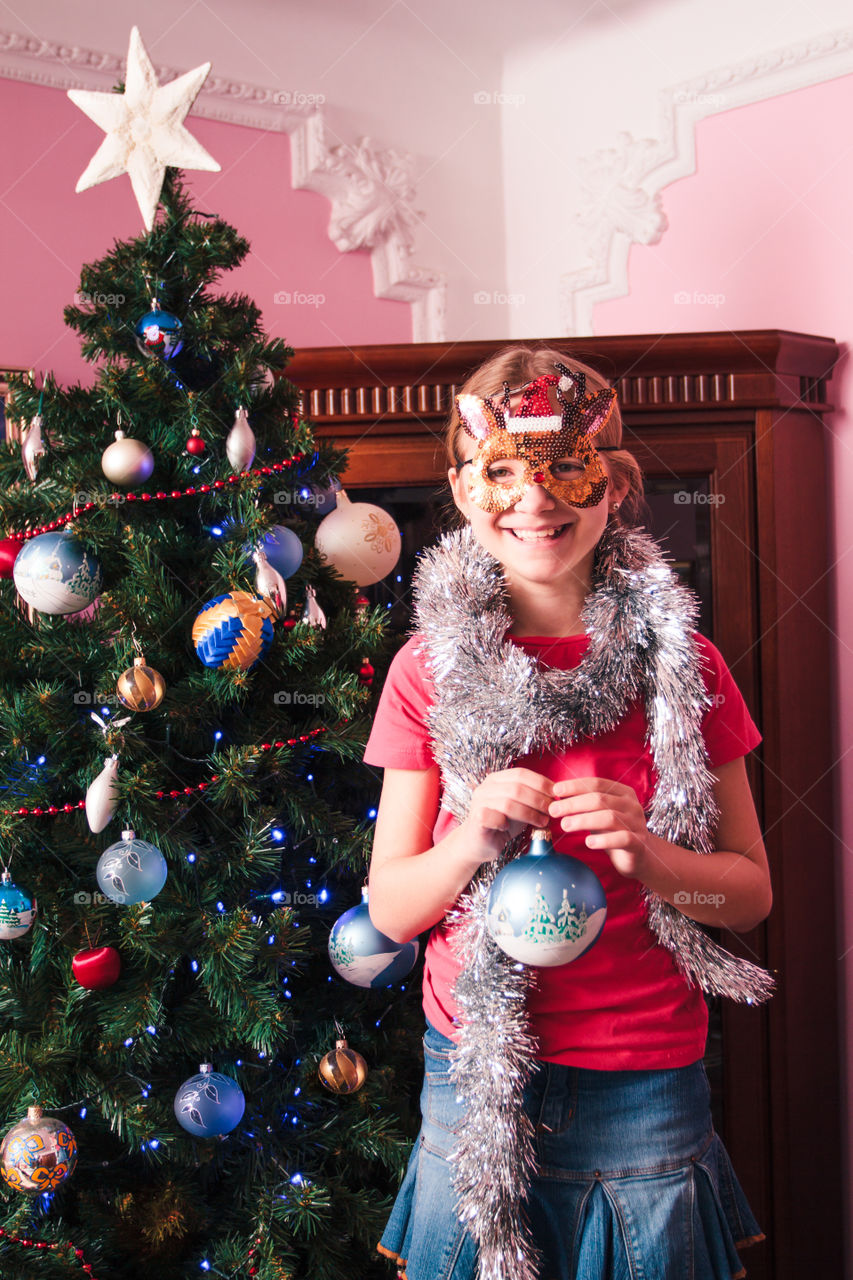 Young girl decorating Christmas tree, holding big Christmas ball. Teenage blonde girl wearing blue jeans skirt and pink blouse and tights. Girl has a funny reindeer mask on her face. Young girl celebrating Christmas at home