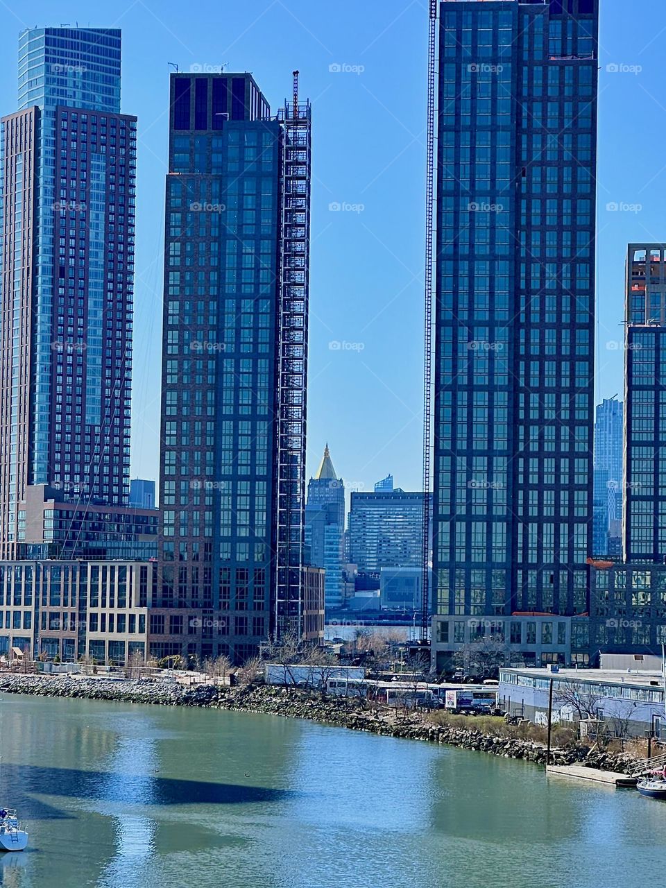 This is „Newtown Creek“ seen from the „Pulaski Bridge“ that connects LIC to „Greenpoint“, Bklyn. Between two recently built LIC high rises we see the „New York Life Bldg“ in „Manhattan“ with the golden triangular roof. 2024. Hypnotic Productions