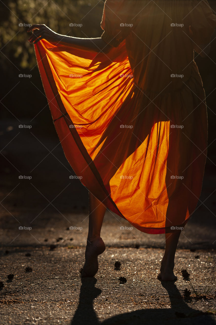 Conceptual dreamy photography of slim young lady posing barefoot while warm sunlight passes through the yellow airy dress, revealing the silhouette of her legs at summer evening