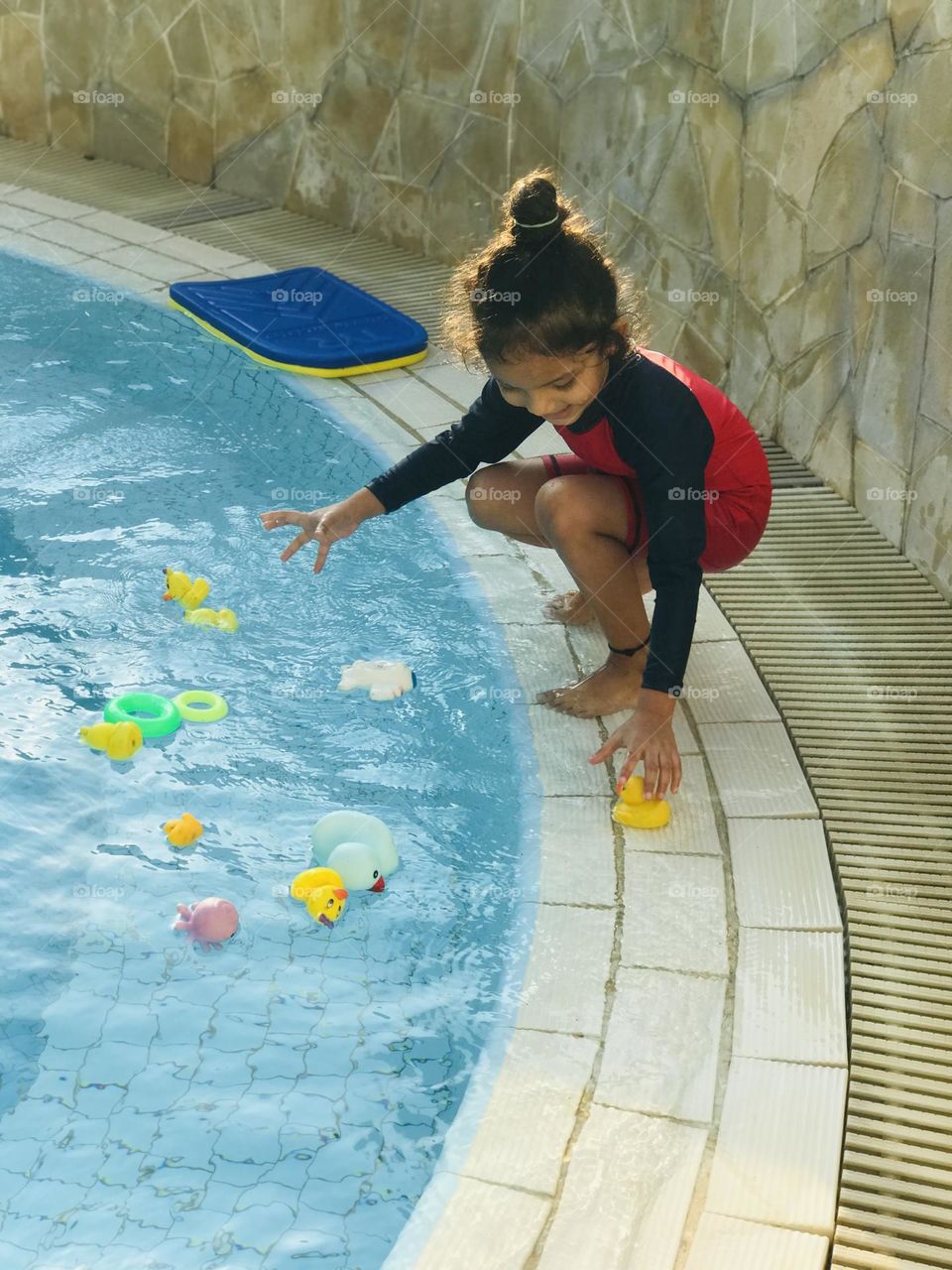 A 4 year old cute girl playing at swimming pool with water toys like ducks,rings etc…and wearing red and black swimsuit.