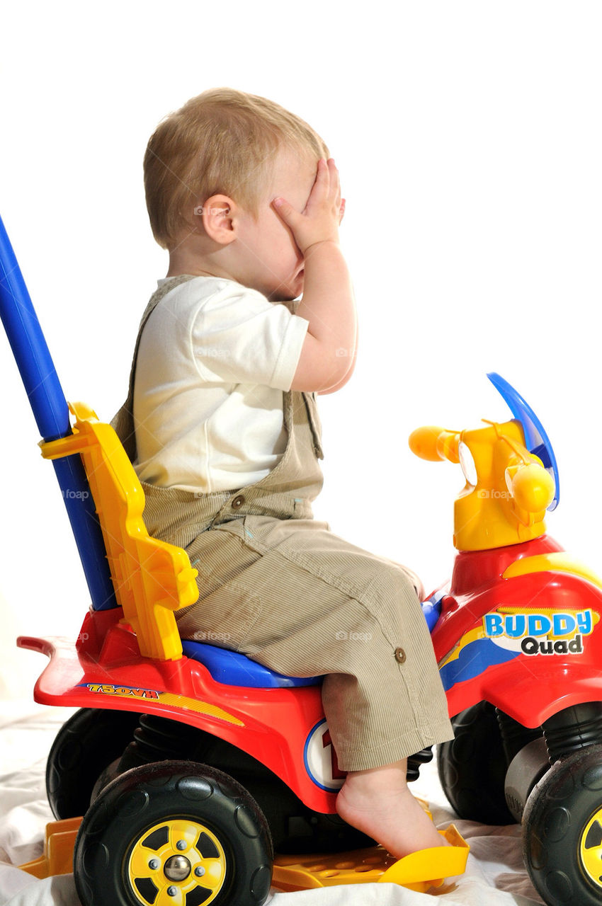 YOUNG WHITE TODDLER BOY SITTING ON A RIDE-ON TOY AND COVERING HIS FACE