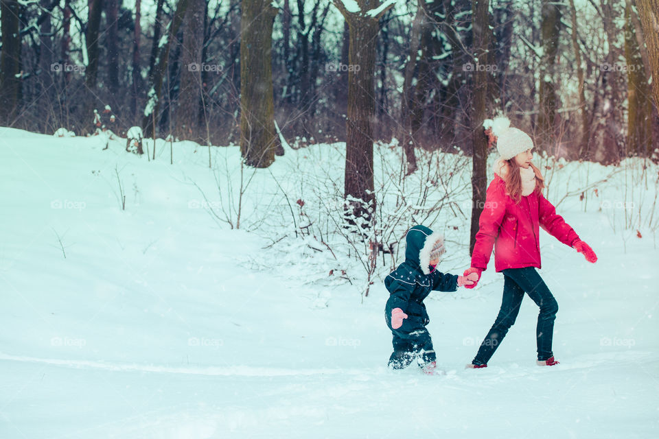 Teenage girl enjoying snow with her little sister. Children are walking through deep snow while snow falling, enjoying wintertime. Sisters spending time together. Girls are wearing winter clothes