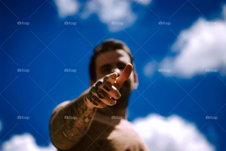 Tattooed man pointing to the camera, with hand and arms tattoos. A beautiful blue sky is on the background.