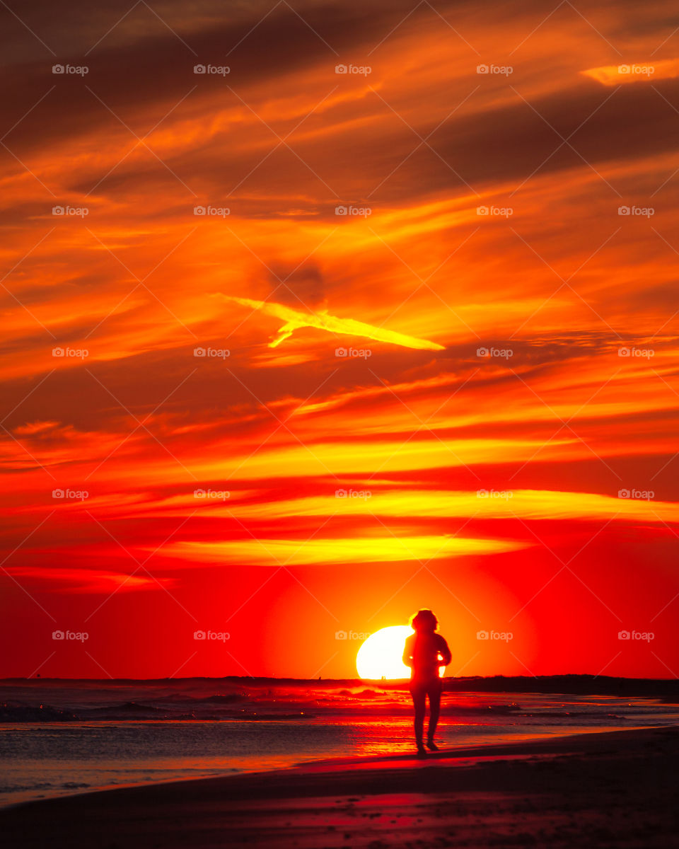 Silhouette of a person walking along the coast, as the sunset creates a vibrant red and orange sky. 