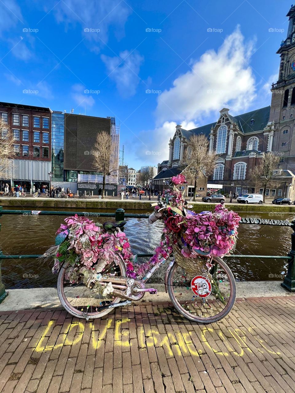 The photo shows a pink bicycle decorated with flowers near a canal in Amsterdam, with the words “LOVE ME” written on the ground, and a historic building in the background.