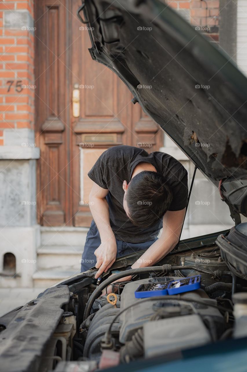 One young caucasian man is unscrewing a burned-out bulb of the front left headlight with a screwdriver in the open hood of a car, standing hunched over on a city street during the day near his house, close-up side view. Car repair concept.
