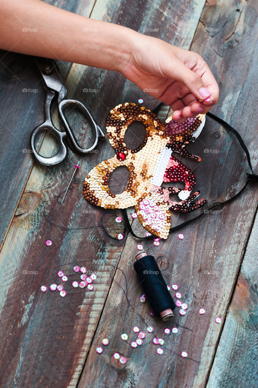 Close shot of female hand decorating Christmas reindeer mask with sequins using thread needle and  scissors following diy ideas on wooden table