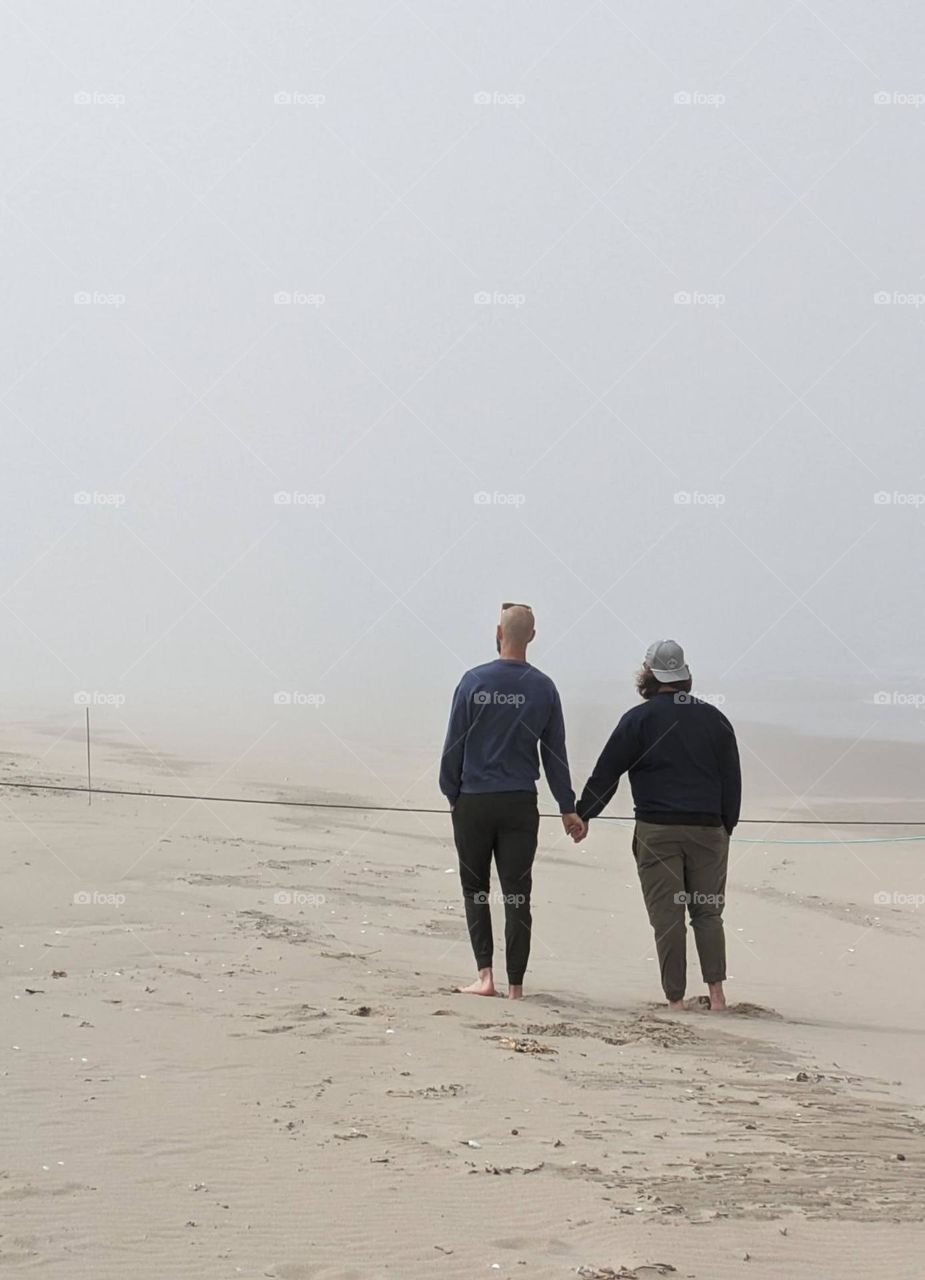 men walking on the beach by the ocean holding hands, foggy beach, men hand holding, partners for life, moody beach vibes