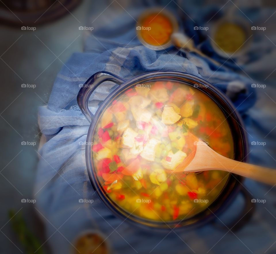 Casserole with freshly cooked chicken soup and a wooden stirring spoon against a dark background.  Food photo, square orientation, top view
