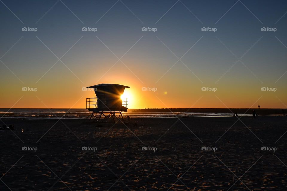 A lifeguard station is silhouetted by the sun setting behind it 