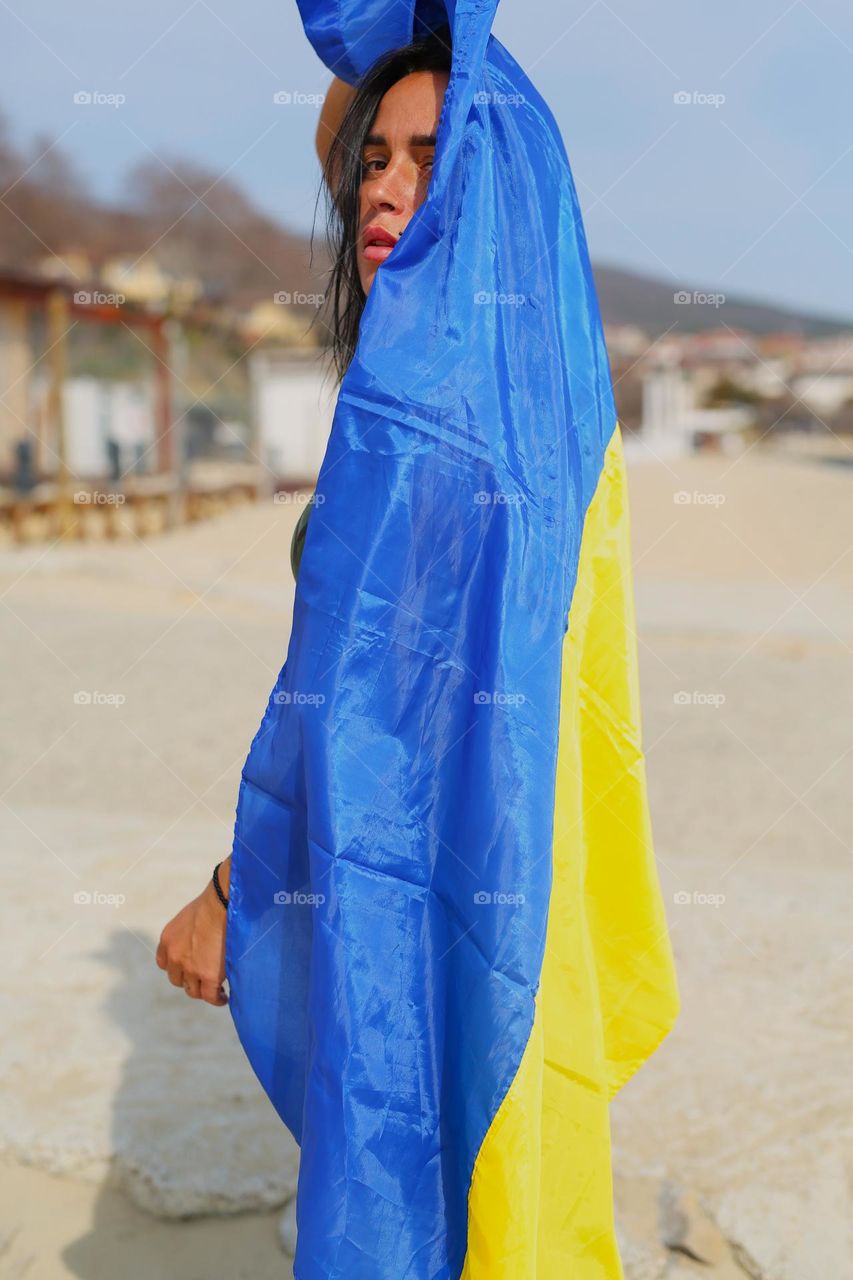 a beautiful girl is wrapped in a flag, yellow and blue, the girl is standing on the seashore, sadness in her eyes