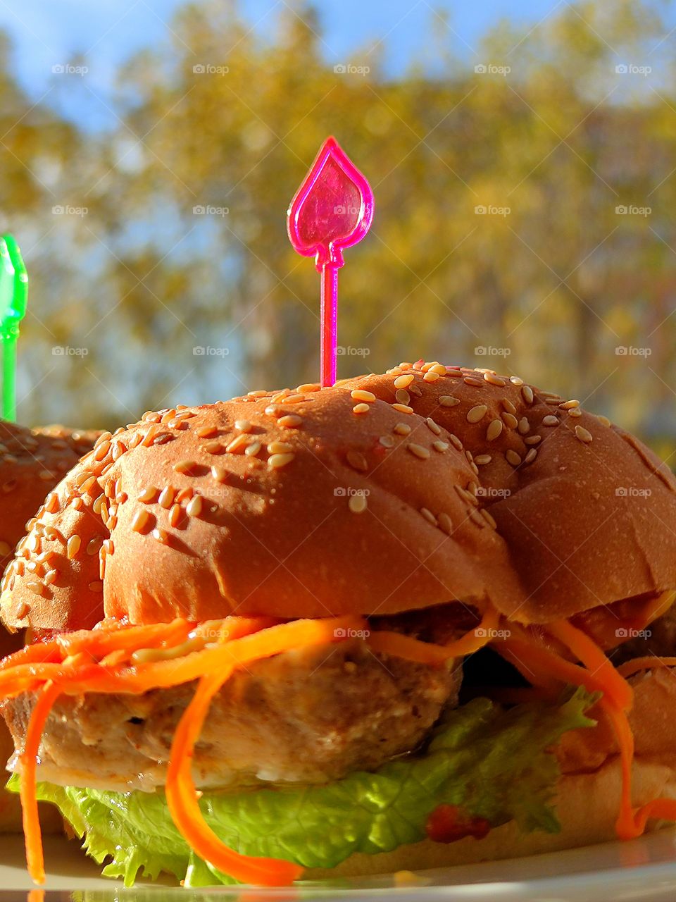 Burger. Sunlight hits the burger and takes on a rich color. Grain bun, inside the is a leaf of green lettuce, mayonnaise, ketchup, cutlet and spicy carrots, which are thinly sliced. The burger is held together by a pink skewer