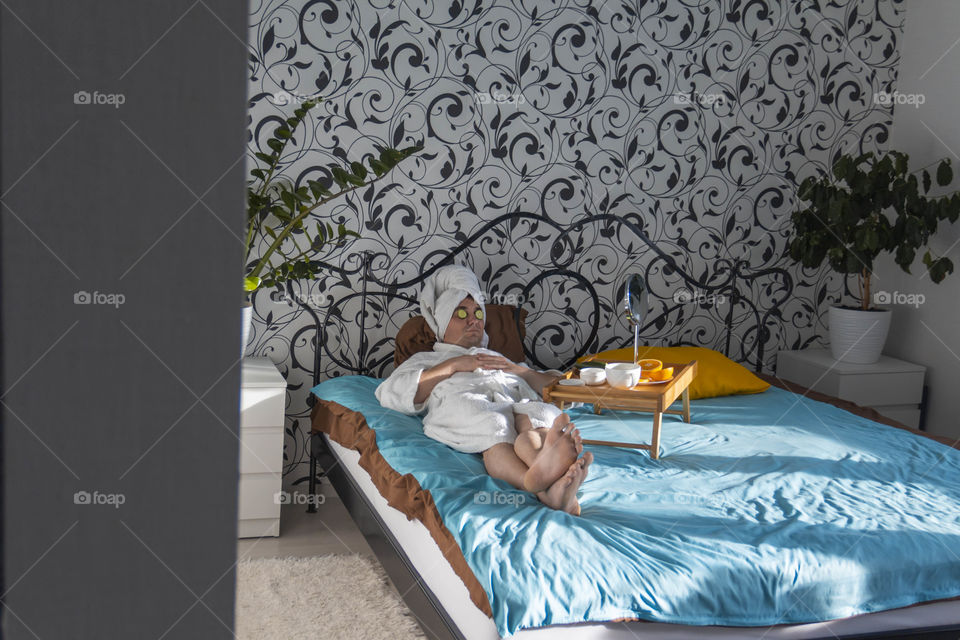 A man and home spa care at home takes care of himself and smears his face with cream, does a manicure at home on a blue background and with oranges and rests.