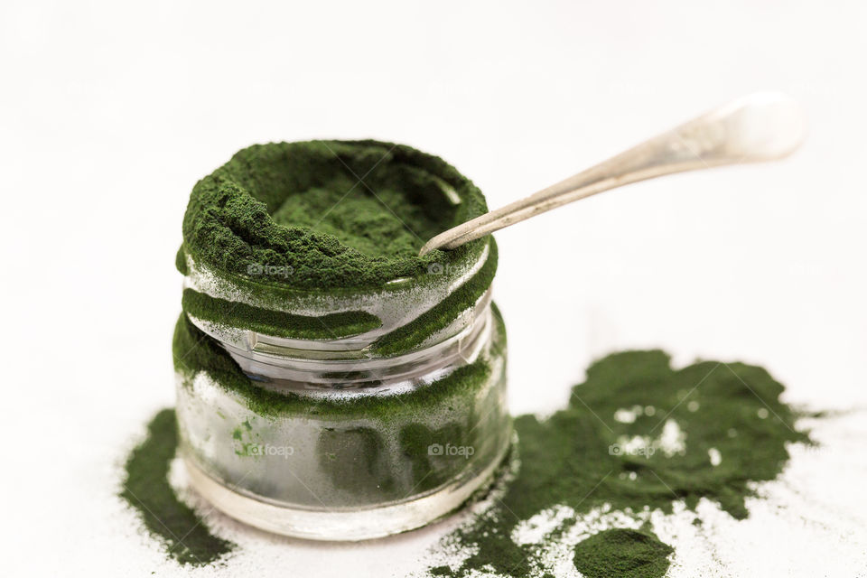 Product healthy green spirulina powder close up in glass jar with spoon on pure white background