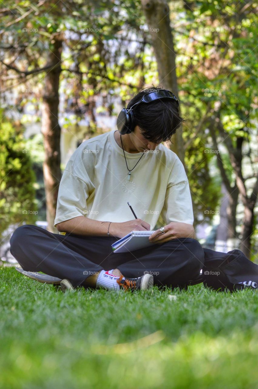 a young man in headphones and loose clothes sits in the park on the lawn and draws in a sketchbook