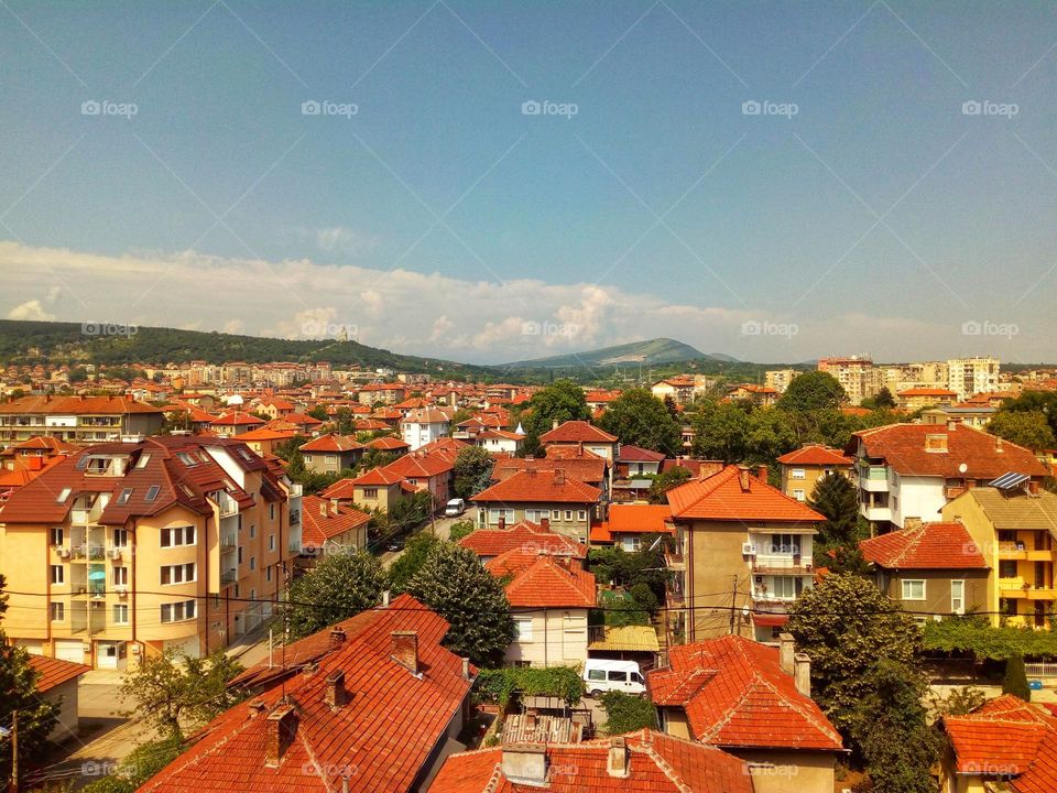 A photo captured on top of a Bulgarian town full of apartment buildings and houses with red tiled roofs and hills in the background