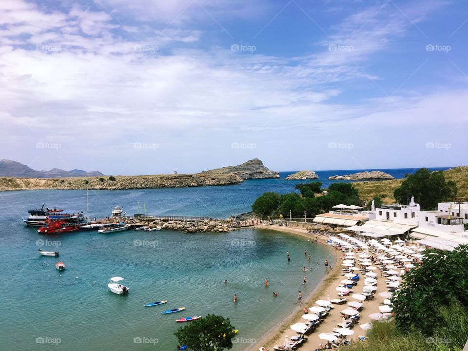 there are many white umbrellas, chairs and a lot of people , playing in cool blue water. There are kayaks, boats and yachts moored on the water, a rocky mountain range and white cloudy blue sky