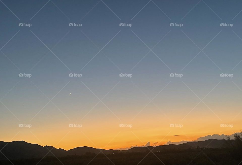 A yellow and orange sunset with mountains and a crescent moon in the background.