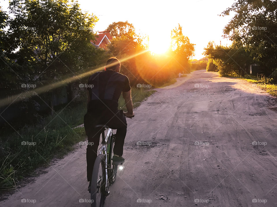 Young man riding a bicycle by the countryside road towards the sunset rays 