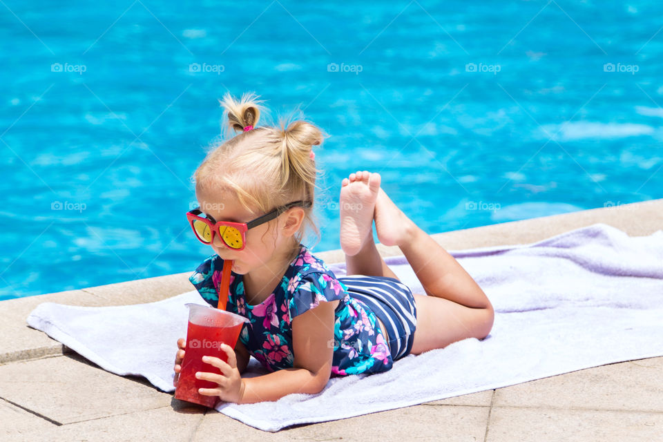 Cute little girl with blonde hair drinking fresh juice near swimming pool 