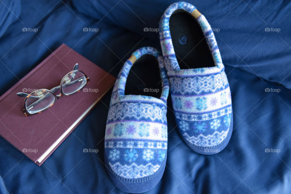 Flat lay of a pair of fleece slippers on a bed next to a book and glasses