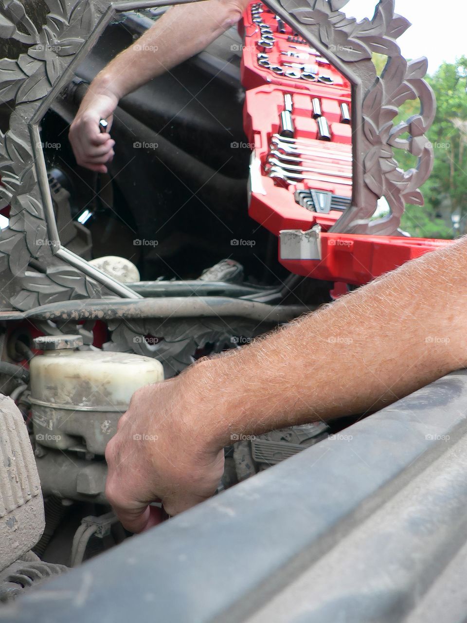 Under the hood of a red pickup truck with combination wrench set tools organized in a red tool hard case by the work station with its reflection from a mirror and the mechanic handyman fixing a piece.