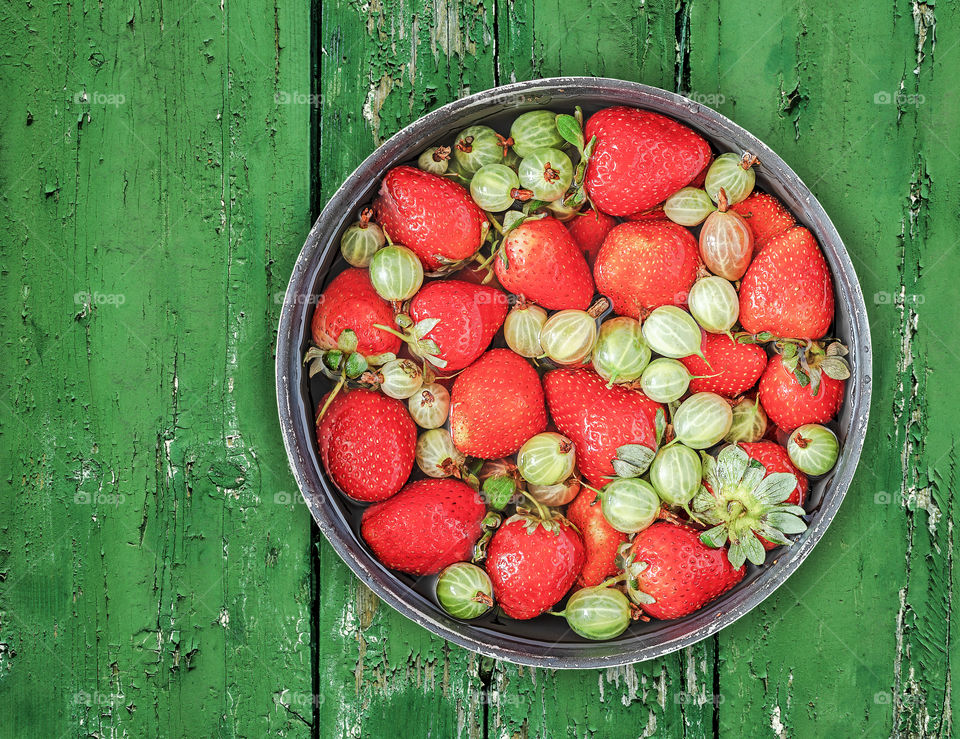 The berry-picking season, the harvest. Fresh natural strawberries and gooseberries in a bowl with water on an old wooden green background. The concept of organic, healthy, seasonal fruits. Food photo