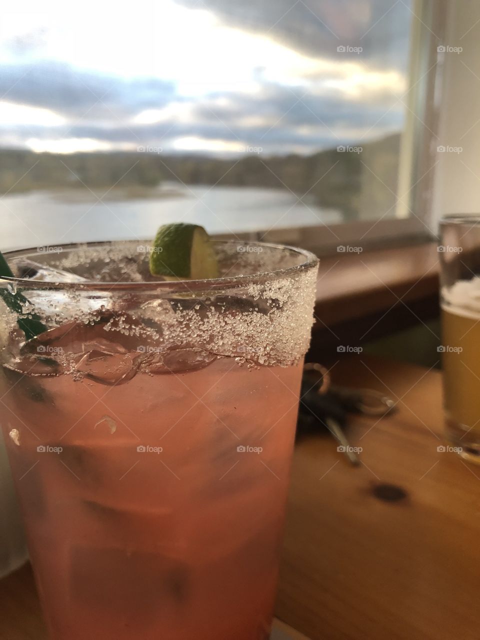 Strawberry Margarita in a clear glass with sugar and lime on rim, a beer in a clear glass, and a set of car keys sitting on a wooden table over looking a window with a view of the water 