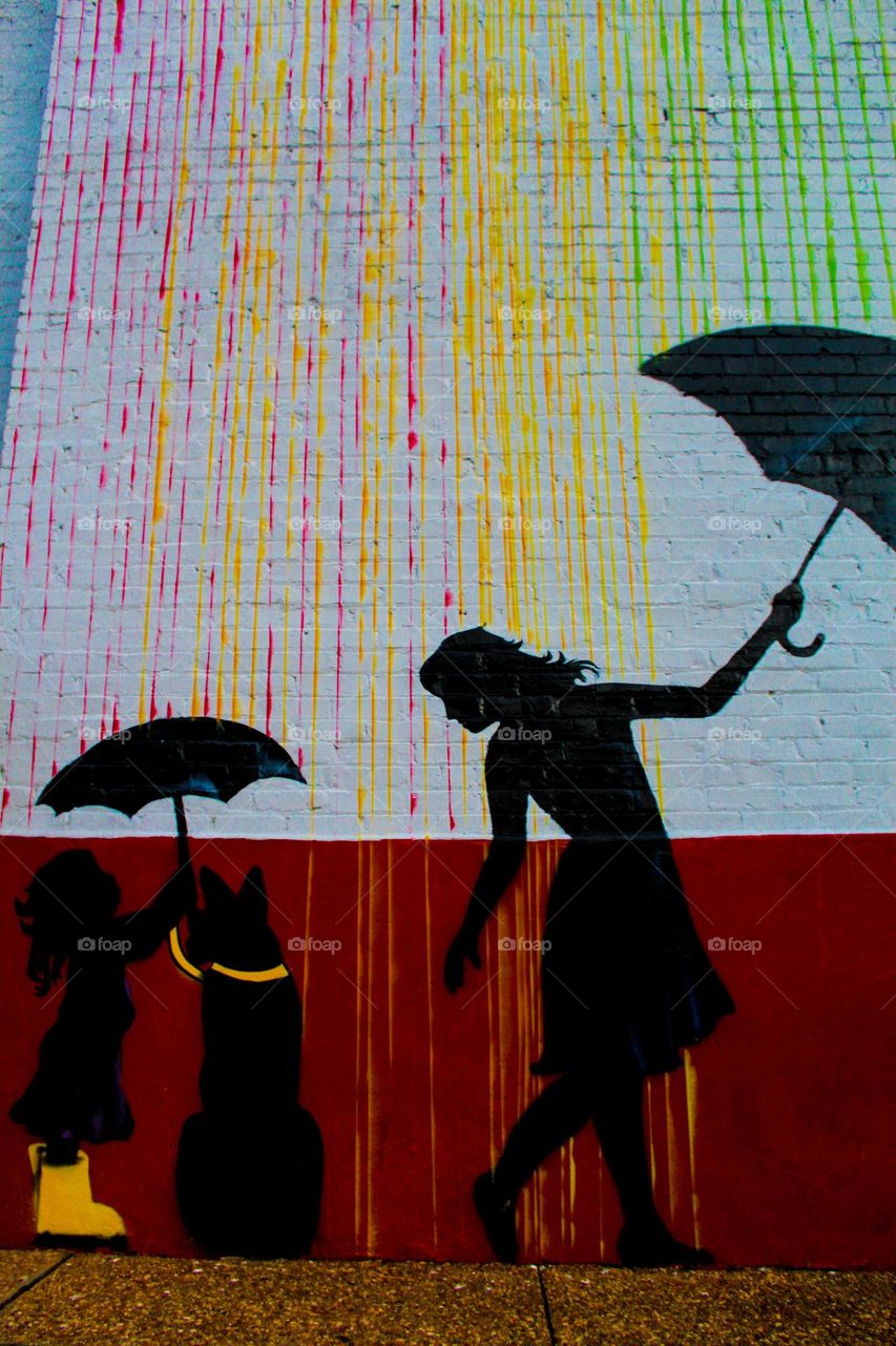 A beautiful piece of artwork by an unnamed artist on the side of a building. Colorful rain covers the woman, and the child shares her umbrella with her dog.