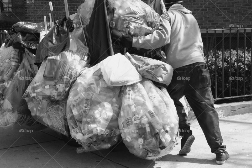 A  back view of a man in Manhattan, New York City pushing carts filled with plastic bags containing  plastic, metal and glass bottles and cans to be recycled for 5 cents per item.