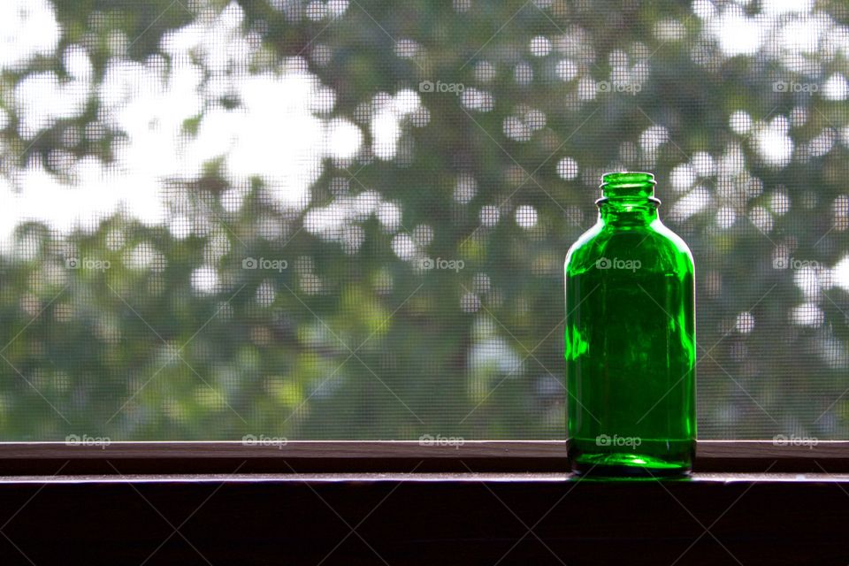 A small, empty green bottle on a windowsill against a blurred tree in the background 